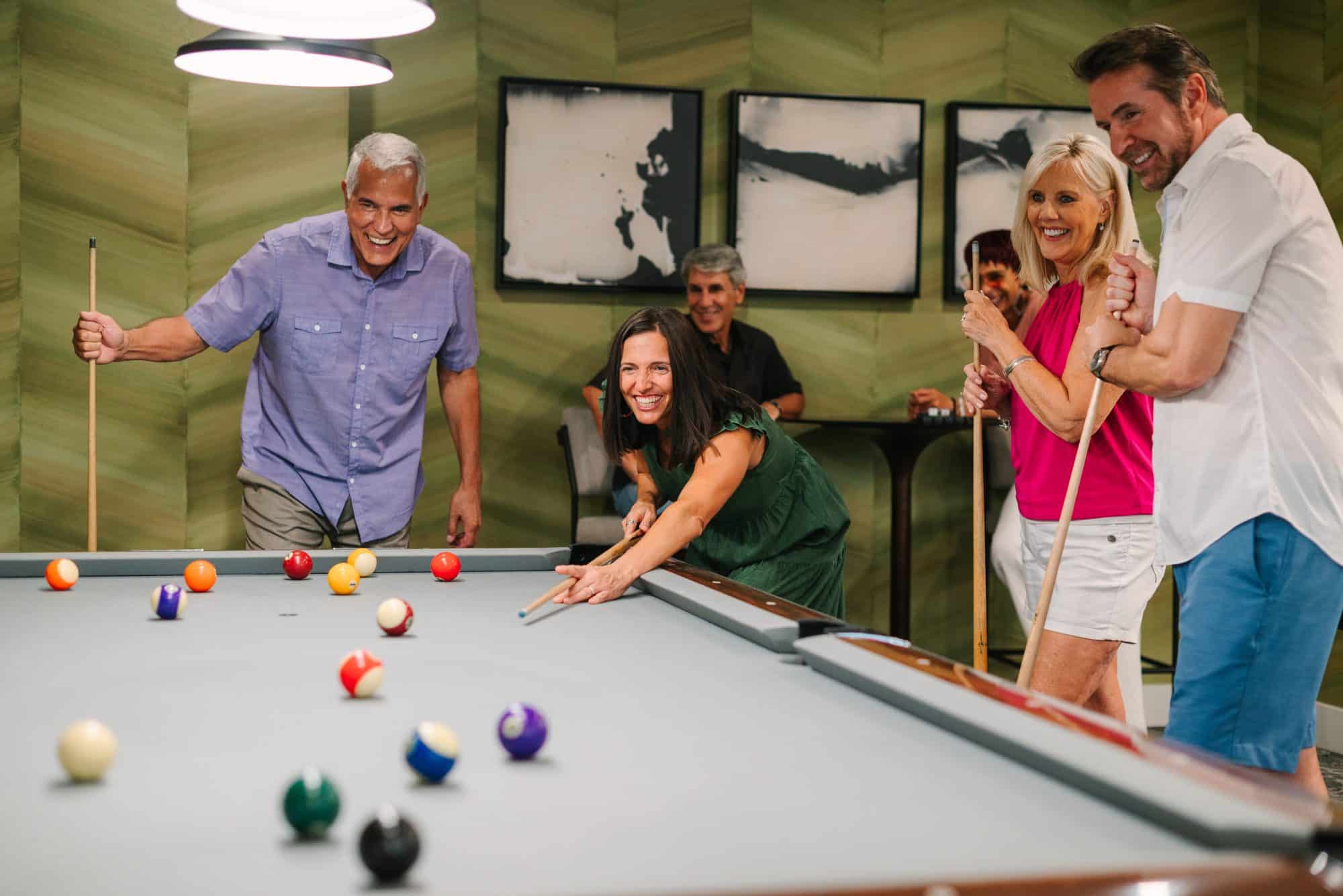 Group of people playing pool in a recreational room, with one woman aiming the cue stick and others laughing and watching in peoria az