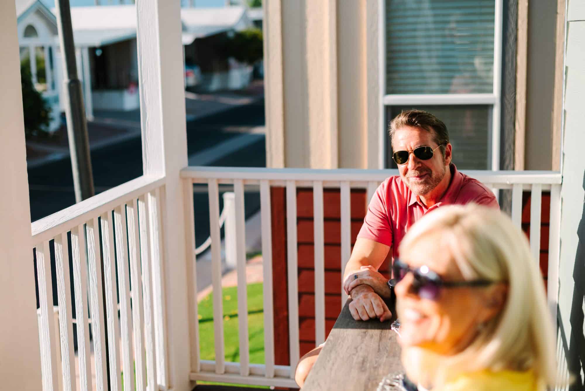 A man with sunglasses and a red shirt sits on a porch table smiling in sunlight, with a woman in the foreground wearing sunglasses and a yellow shirt in peoria az